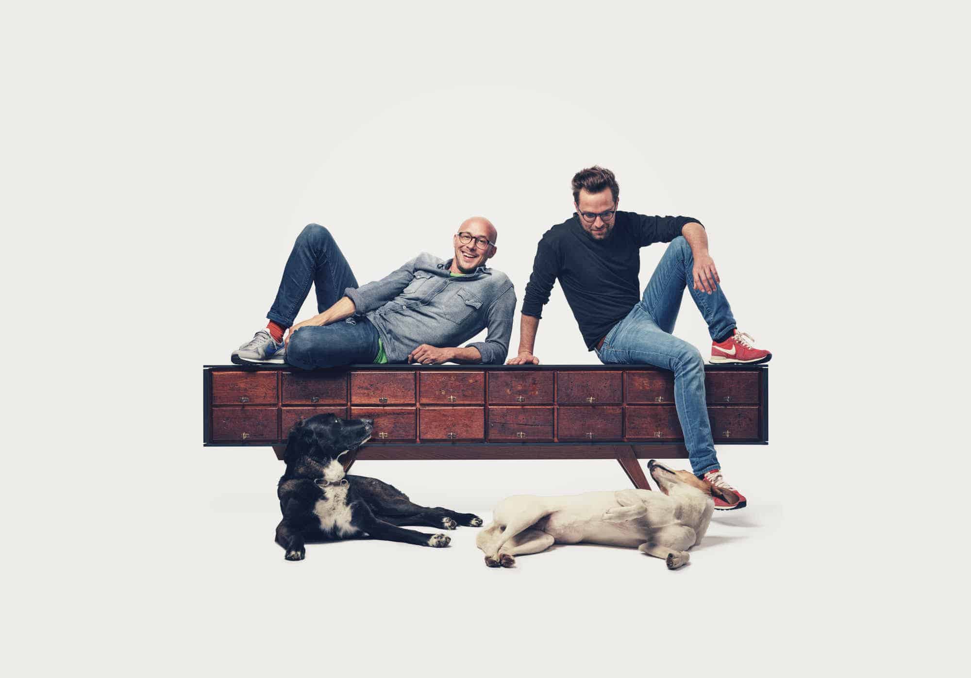 Alexander Mompo und Philipp Egenter sitzen auf einem Sideboard, fotografiert im Studio mit den zwei Hunden vorne dran