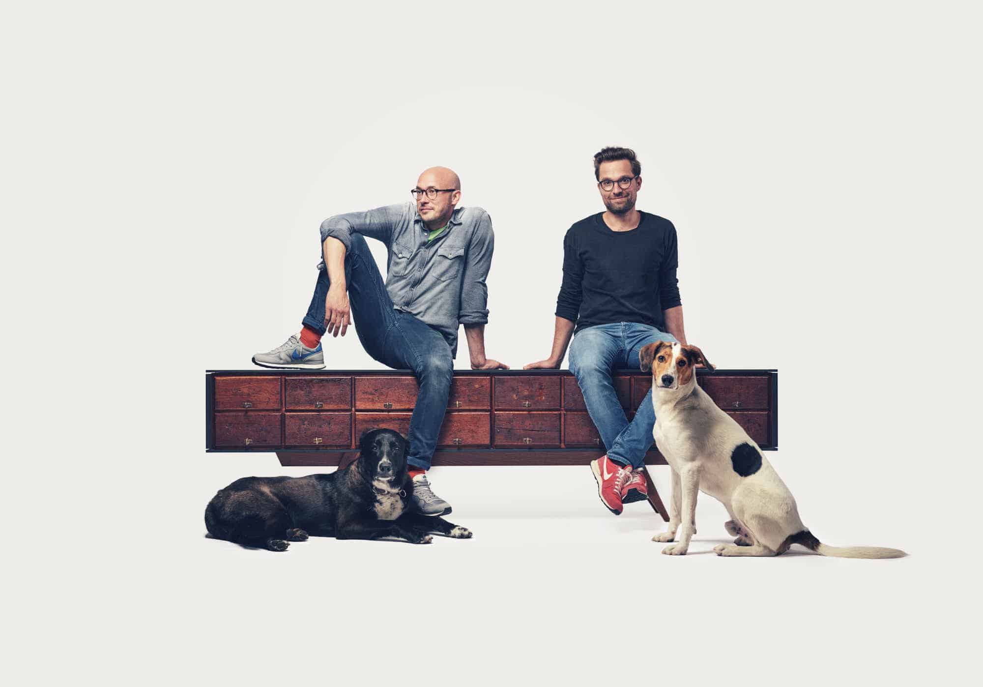 Alexander Mompo und Philipp Egenter sitzen auf einem Sideboard, fotografiert im Studio mit den zwei Hunden vorne dran