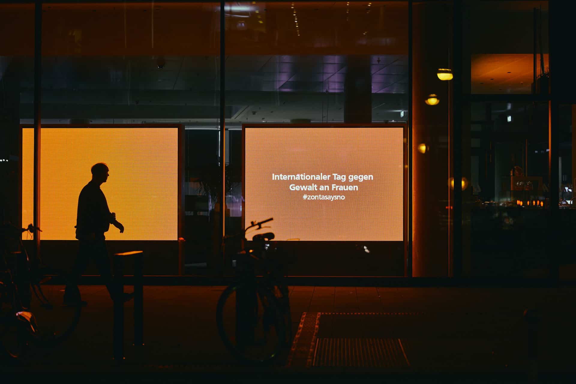 Mann geht vor einem Schaufenster bei Nacht. Displays sind orange und auf einem Steht: Internationaler Tag gegen Gewalt an Frauen