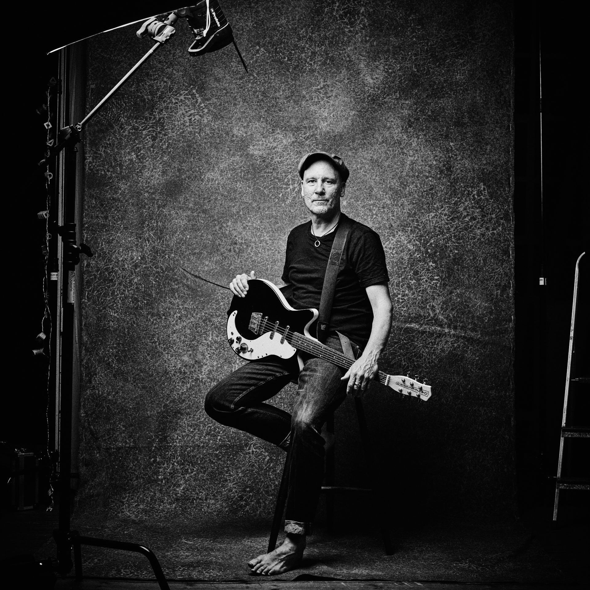 Schwarz-Weiß Aufnahme mit Sascha Bendiks in meinen Fotostudio. Er sitzt auf einen handgemachten Barhocker, eine Schwarze Gitarre auf dem Schoss. Blick in die Kamera. Der Blitz und das Stativ sind mit auf dem Bild.