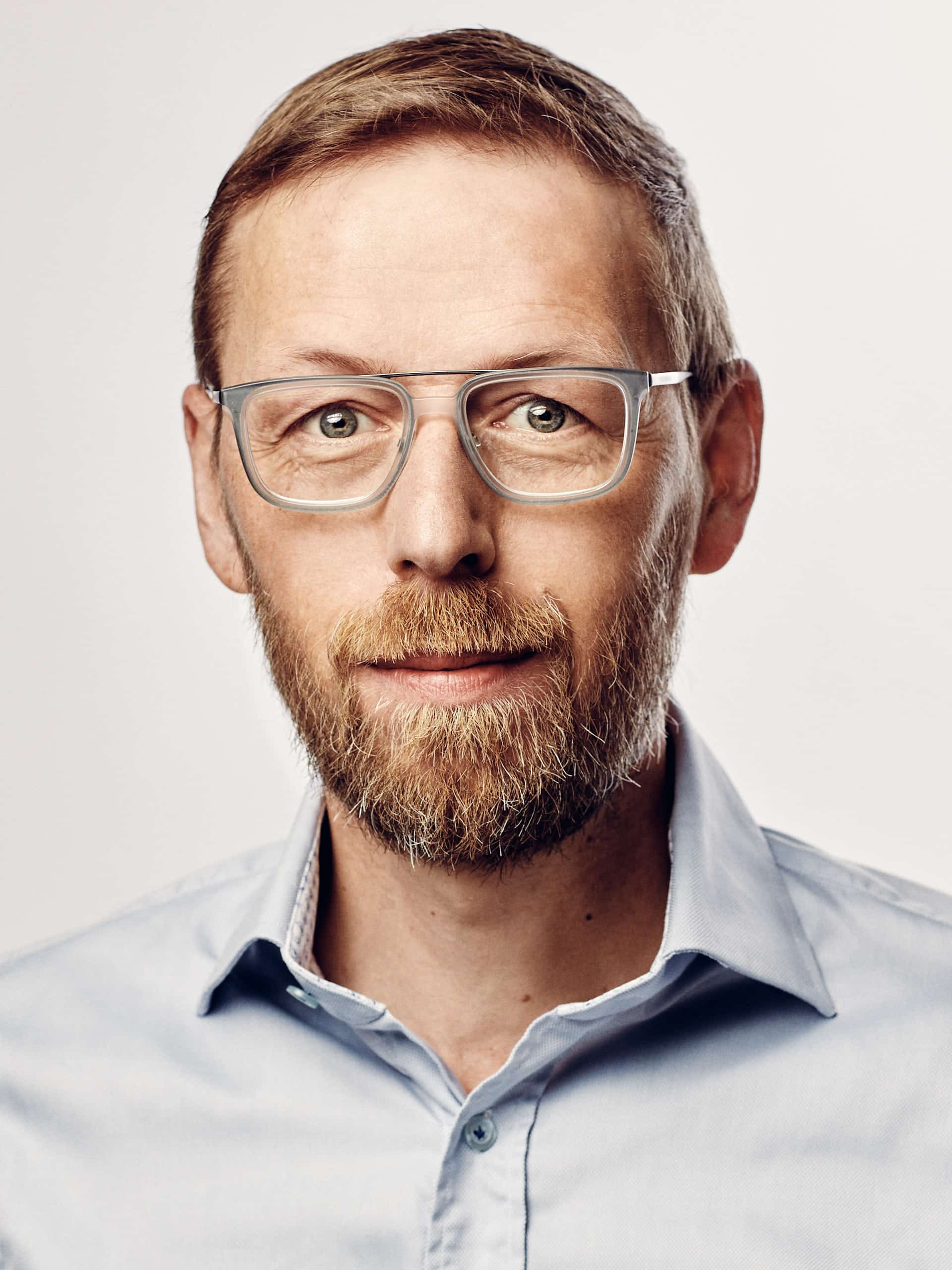 Close up Portrait mit dem Frisör Günter Ducke im Studio vor hellen Hintergrund. Er hat eine Brille auf und schaut direkt in die Kamera.