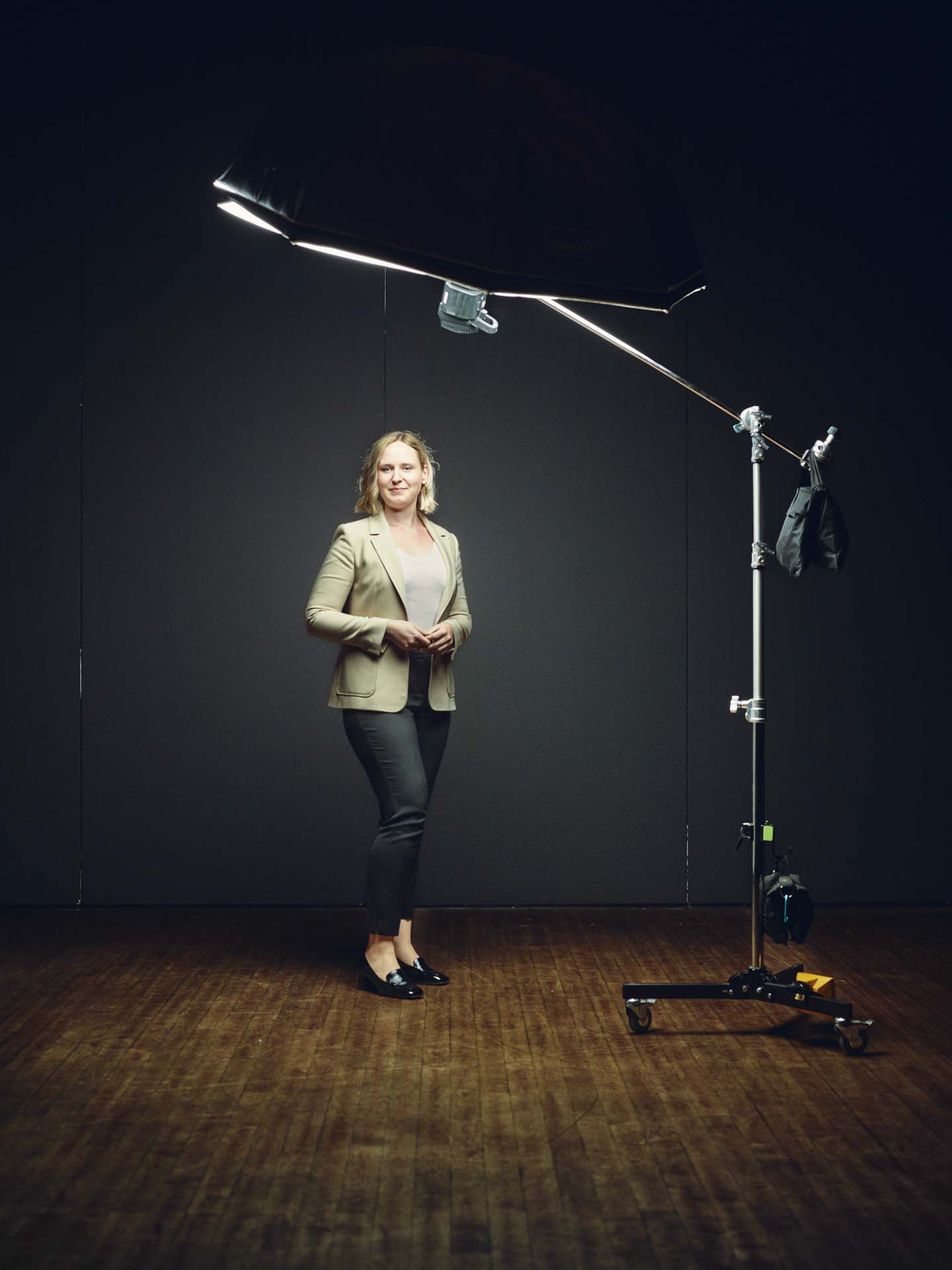 Studio-Portrait von Sarah Schüber, sitzend auf einem Stuhl unter einem großen Softlight. Die Aufnahme zeigt die Lichtquelle und den Aufbau, wodurch ein Blick hinter die Kulissen entsteht. Fotografiert von Felix Groteloh.